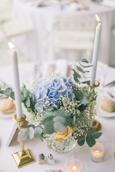 a centerpiece with blue flowers and greenery sits on a white tablecloth surrounded by candles