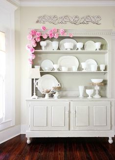 a white china cabinet with pink flowers on the top and dishes on it's shelves