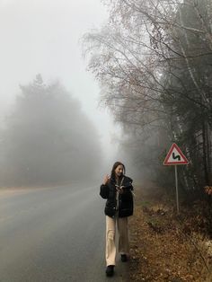 a woman standing on the side of a foggy road with trees in the background