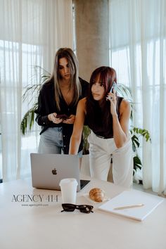 two women standing next to each other in front of a laptop computer and cell phone