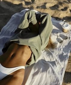 a woman laying on top of a white towel next to a cup and bottle in the sand