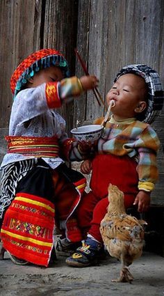 two young children eating out of bowls while standing next to a wooden fence and chicken