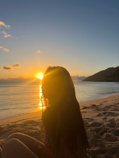 a woman sitting on top of a sandy beach next to the ocean at sun set