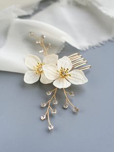 two white flowers are attached to a gold hair comb on a gray surface with a white scarf in the background