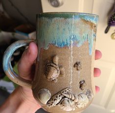 a hand holding a coffee mug with sand and shells on it