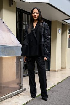 a woman standing on the sidewalk in front of a building wearing a black suit and heels