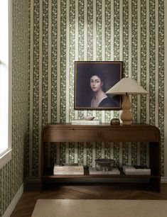 a table with a lamp on it in front of a wallpapered room that has a portrait of a woman