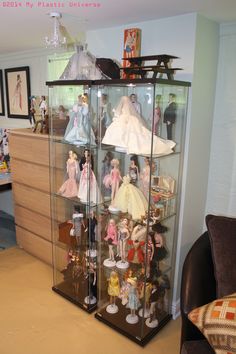 a display case filled with lots of dolls on top of a wooden dresser next to a chair