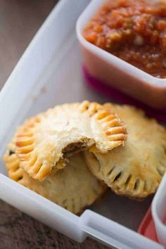 two small pastries in a plastic container with dipping sauce on the side and another dish