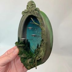 a hand holding up a miniature clock with trees and mountains on it's face