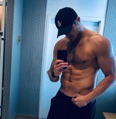 a shirtless man taking a selfie in the mirror with his cell phone and hat on