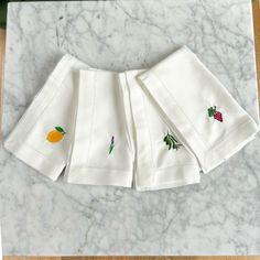 four white napkins with fruit embroidered on them sitting on a marble counter top next to a knife