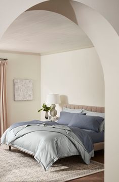 a bed with blue sheets and pillows in a white room next to a lamp on a table