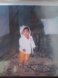 an old photo of a young child in a white robe and orange pants standing on a rug