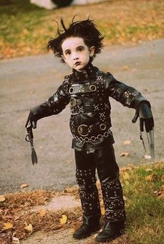 a young child dressed in black leather clothes and holding a pair of scissors, standing on the sidewalk