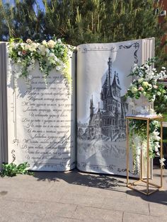 an open book sitting on top of a sidewalk next to flowers and greenery in front of a building