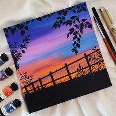 some paint and brushes are laying on a bed next to an easel with a sunset in the background