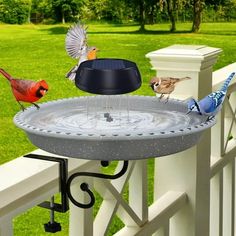 three birds are drinking water from a bird bath on a white fence with green grass and trees in the background