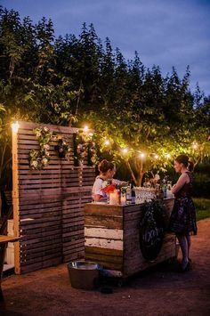 two people standing at an outdoor bar with lights on the trees in the back ground