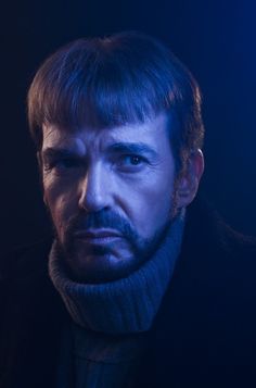 a man with dark hair and beard wearing a black coat looking at the camera while standing in front of a blue light