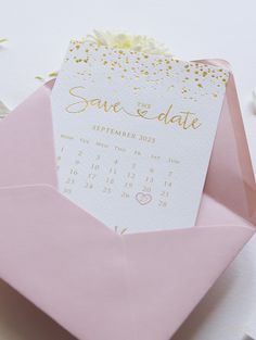 a pink envelope with a white and gold save the date card in it next to some flowers