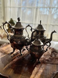 two silver teapots sitting on top of a wooden table next to a window