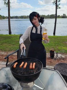 a woman in an apron is cooking hot dogs on a grill with a bottle of beer