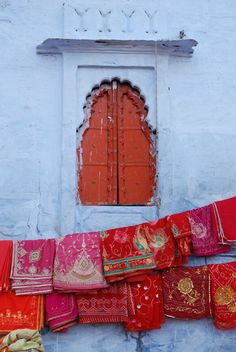 an old building with many different colored fabrics hanging from it's side wall and a red door in the background