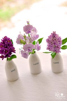 three white vases with purple flowers in them on a tableclothed surface, one has the word love written on it