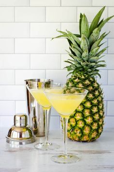 two glasses filled with yellow liquid next to a pineapple on a counter top and silver shakers