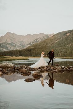 a bride and groom are walking along the shore of a mountain lake with their reflection in the water