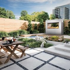 an outdoor dining area with table, benches and fire place in the center surrounded by greenery