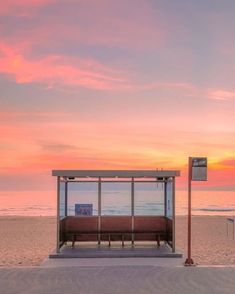 a bench sitting on top of a sandy beach under a pink and blue sky with the ocean in the background