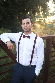 Groomsman wearing a white button up shirt with a dark burgundy bow tie and suspenders. Burgundy Ring Bearer Outfit, Navy And Burgundy Wedding Groomsmen Jeans, Flower Boy Burgundy, Suspender Outfits For Boys, Maroon Suspenders And Bow Tie, Maroon Bow Tie, Red Bow Tie And Suspenders, Chambelanes Outfits, Grooms Suits