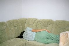 a woman laying on top of a green couch next to a white wall and cat