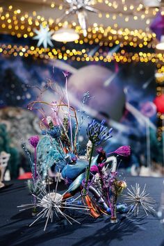 an arrangement of flowers and plants on a table in front of a wall with lights