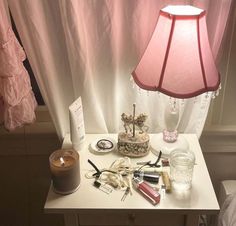 a table topped with lots of different items next to a pink lamp and window curtain