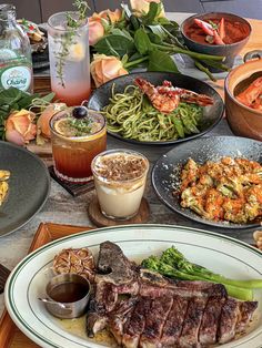 a table full of food including steak, pasta and other dishes with drinks on it