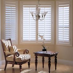 a living room with two chairs and a table in front of three windows that have white shutters on them