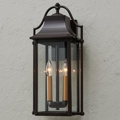 an outdoor light with three candles lit on it's side against a white wall