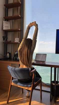 a woman sitting at a desk in front of a window with her arms up to the sky