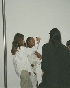 three women standing next to each other in front of a white wall and one woman is holding her hand up