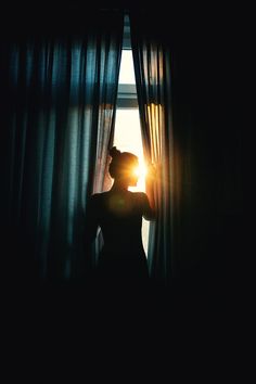 a woman standing in front of a window with the sun shining through her sheer curtains