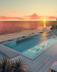 an outdoor swimming pool with lounge chairs and the sun setting over the ocean in the background