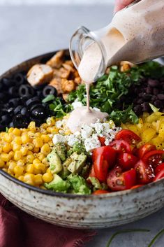 someone pouring dressing over a salad in a bowl with corn, tomatoes, black olives, lettuce and other vegetables