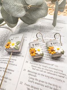 two small flower earrings sitting on top of an open book next to a green plant