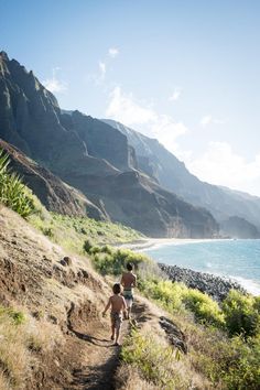 two people walking down a trail near the ocean with mountains in the backgroud