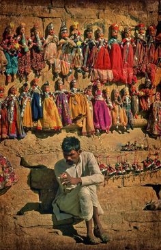 a man sitting on top of a rock next to a wall covered in lots of dolls