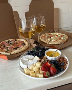 two pizzas, cheese and fruit on a table with wine glasses in the background