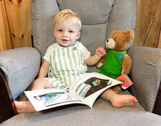 a little boy sitting in a chair with a teddy bear and book on his lap